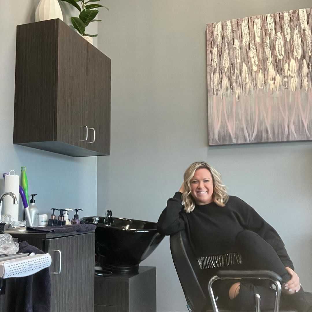 Woman in a salon, sitting on a chair, smiling with a sink and hair products in the background.