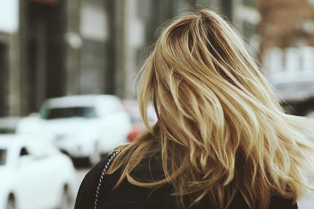 Rear view of a person with long, blonde hair walking in an urban setting with cars in the background.